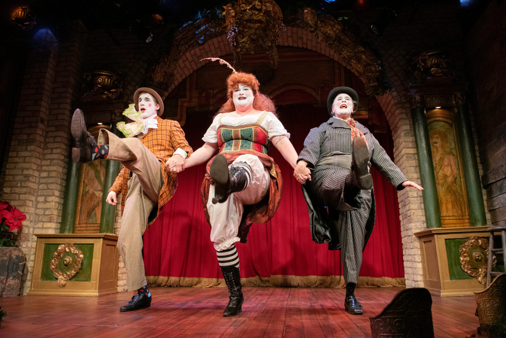 Three costumed charaters kick in unison on a theatre stage.