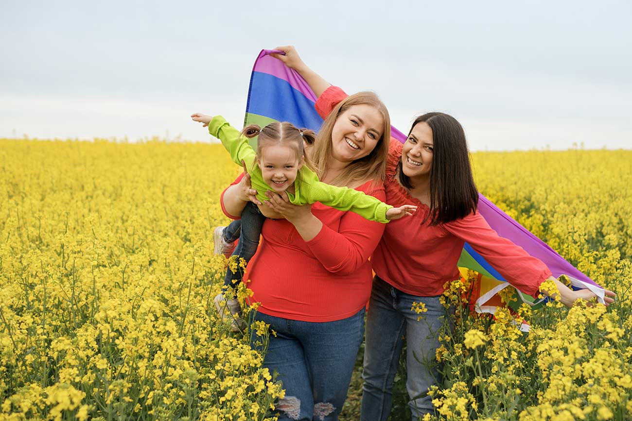 A young couple with a small child and an LGBT flag in a field with yellow rapeseed flowers are laughing merrily.