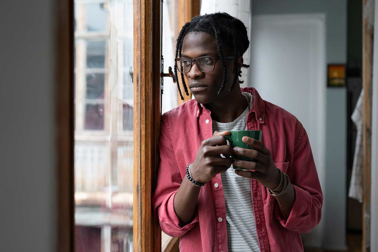 Lonely sad African man looking out window standing indoors suffering due to separation.