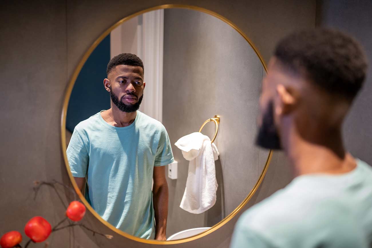 Unhappy African American man looking in mirror, standing in bathroom at home, sad.