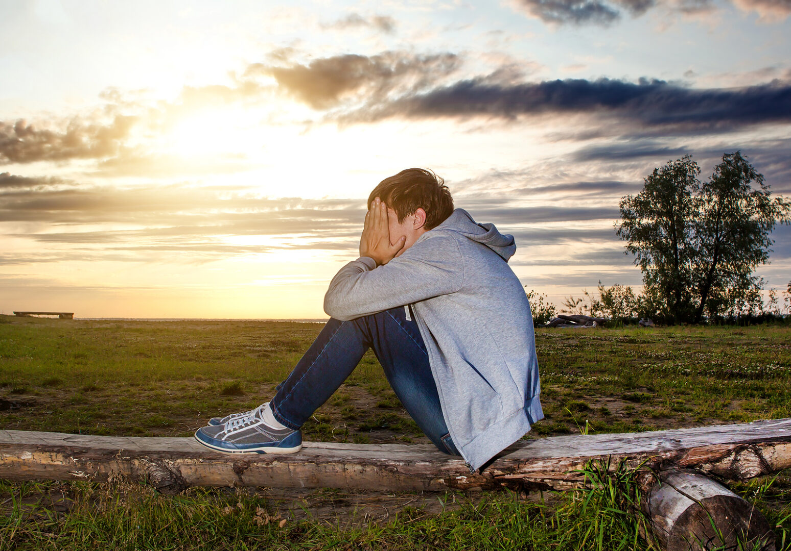 Lonely teenage boy sitting alone on a log in the sunset.
