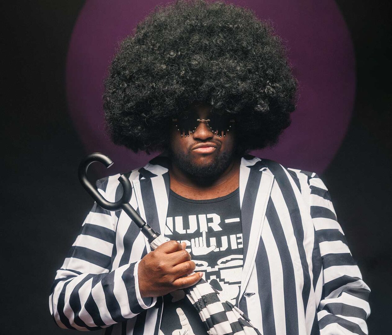 Promo shot of Nur-D in a white and black stripe suit with puffy afro wig.