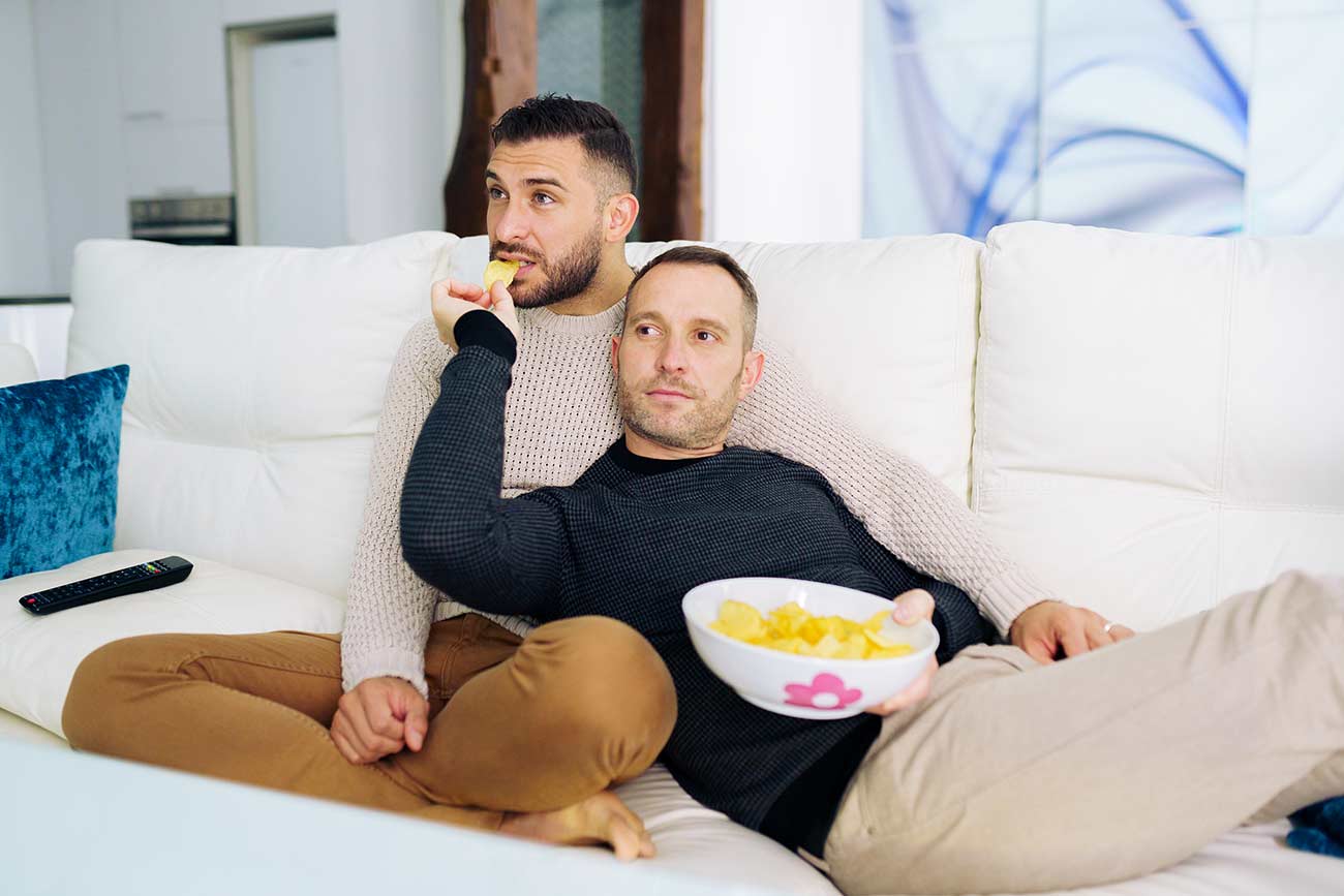 Gay couple sitting on the couch at home watching something on TV and having a snack.